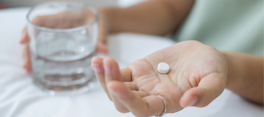 Close-up on woman's hands. One holds a small white pill and the other holds a glass of water.