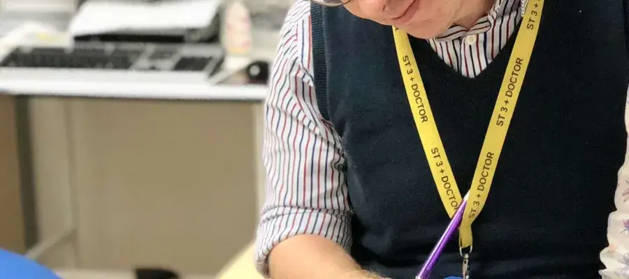 'Neil Ryan is standing in an office with a computer in the background. He is looking and pointing at an image in a textbook called "Modern Pathology". He is wearing a striped collared shirt and a blue sleevless jumper'