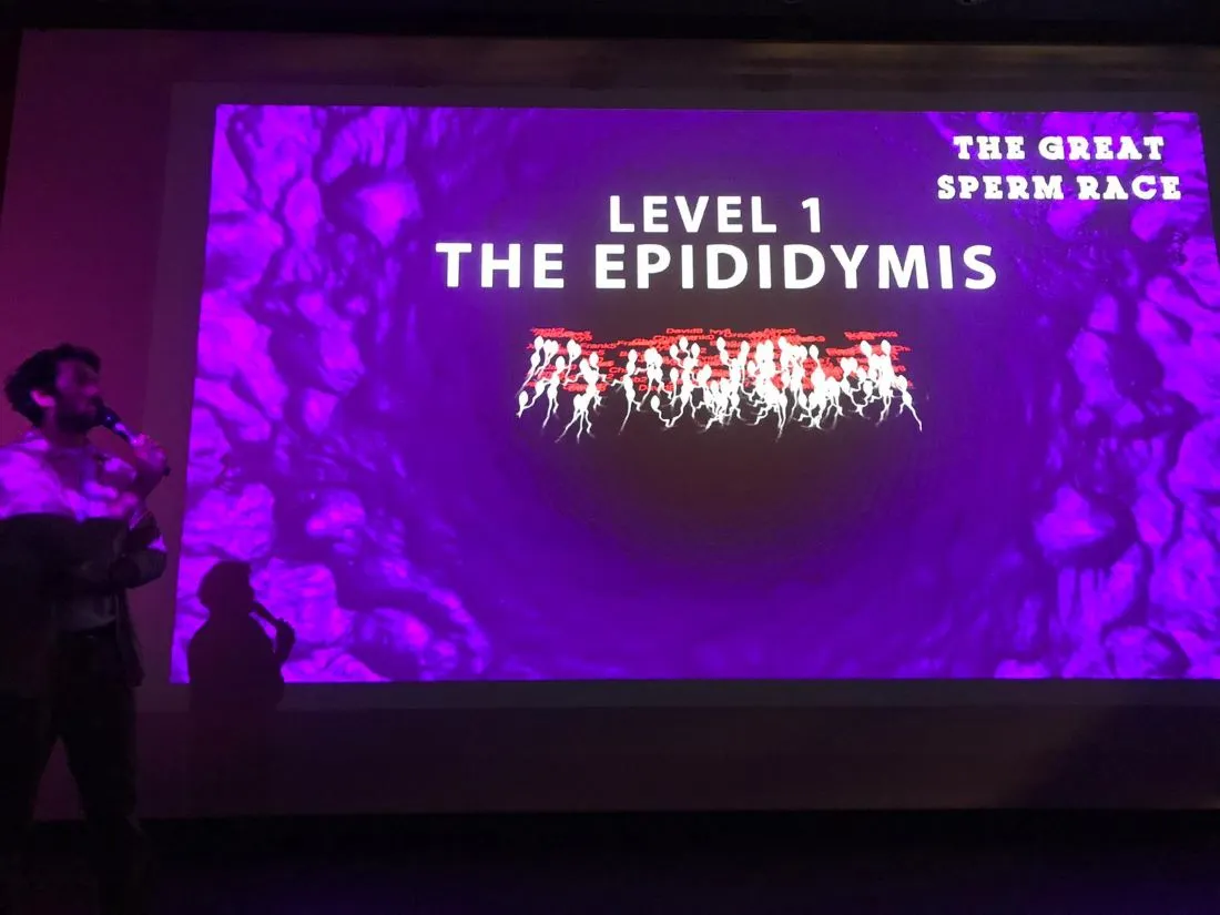 Toby Peach in a dark room with microphone onlooks large purple prjection showing 100 white sperm. 'Level 1 the epedidymis'. 