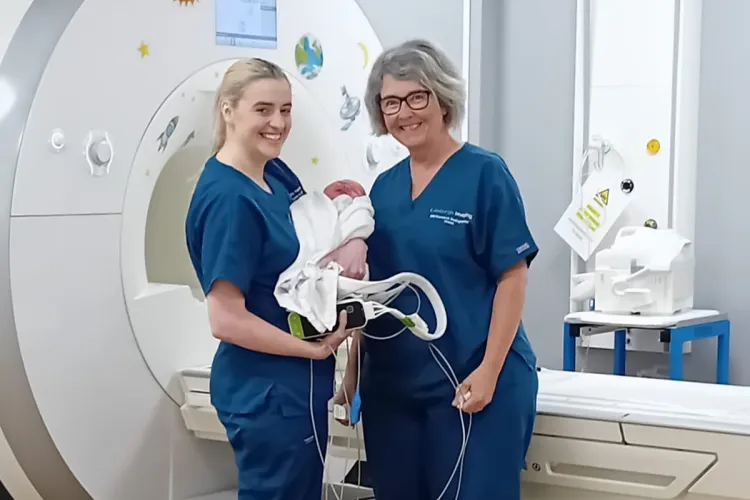 Two clinicians smiling with baby infront of MRI machine