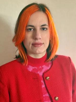 Rocio has bright orange hair and a red cardigan and is smiling at the camera