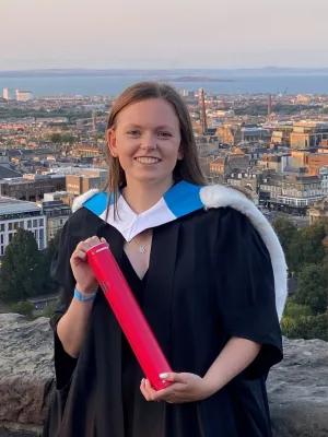 Laura Edwards is wearing a graduation gown, with the Edinburgh skyline in the background and holding a red diploma tube